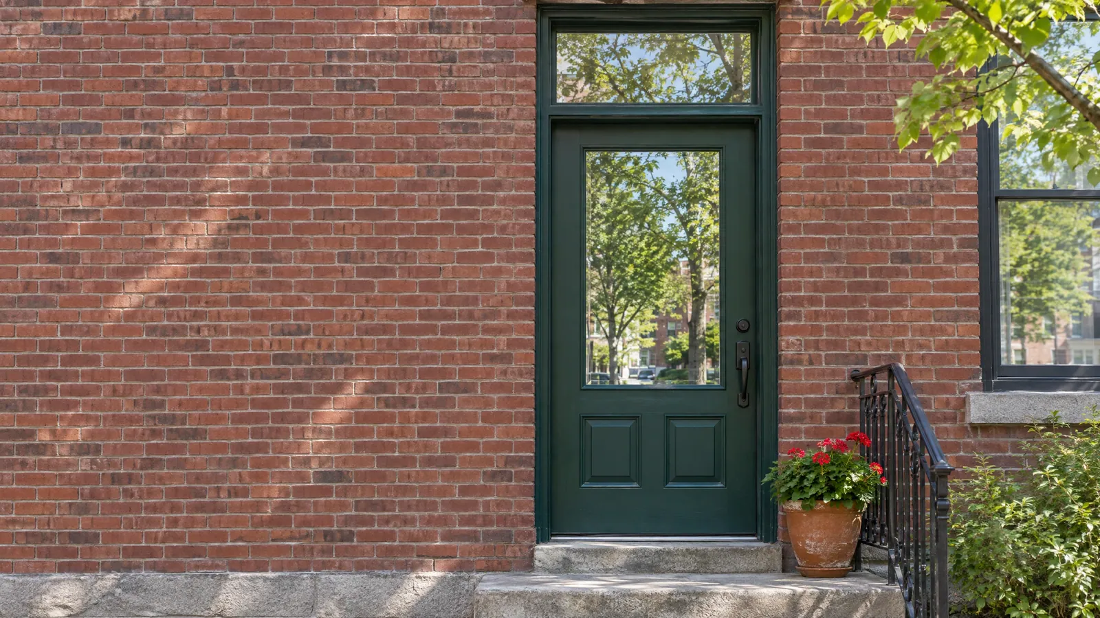 Dark green wood entry door in a brick plex wall, with a tall glass panel in the door, a clear transom above it and geraniums on the stone step